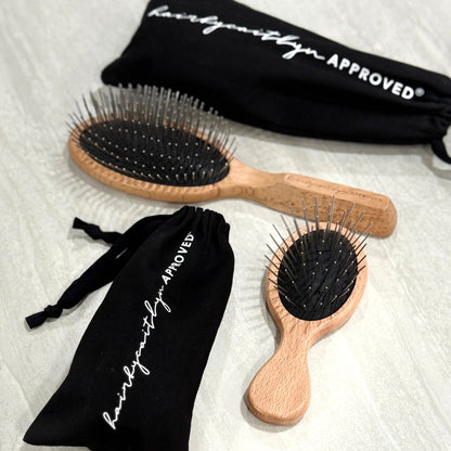 Two wooden hairbrushes with rounded metal pins on a black cushion, accompanied by black drawstring bags with 'HAIRBYCAITLYN APPROVED' text on a travertine surface.