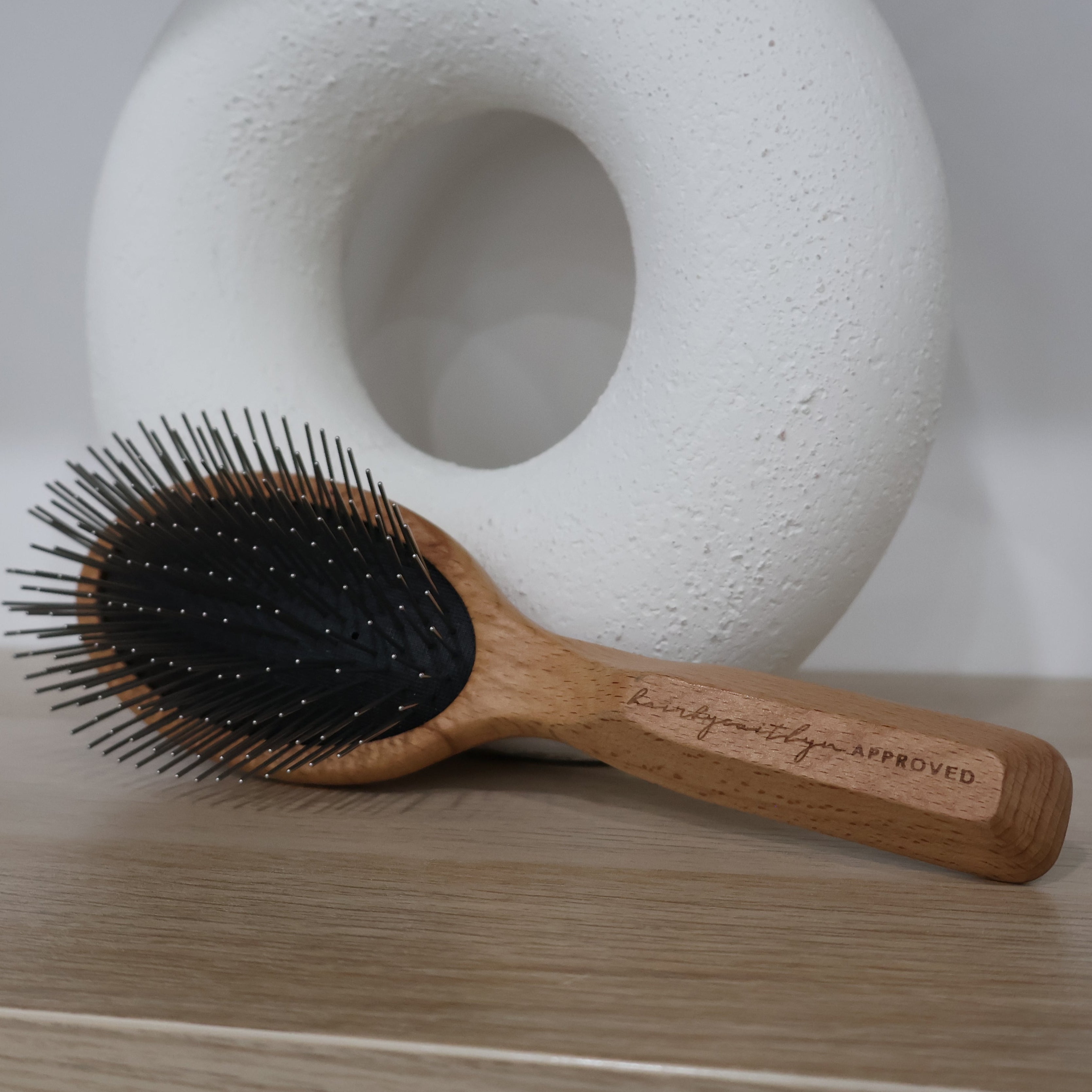 Wooden hairbrush with metal pin bristles on a wooden surface with a white vase in the background
