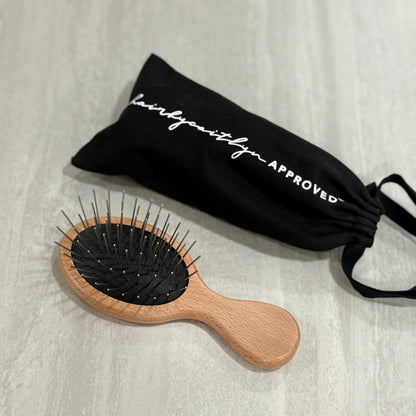 Wooden hairbrush with metal pin bristles and a black dust bag labeled 'hairbycaitlyn APPROVED® on a light gray background.