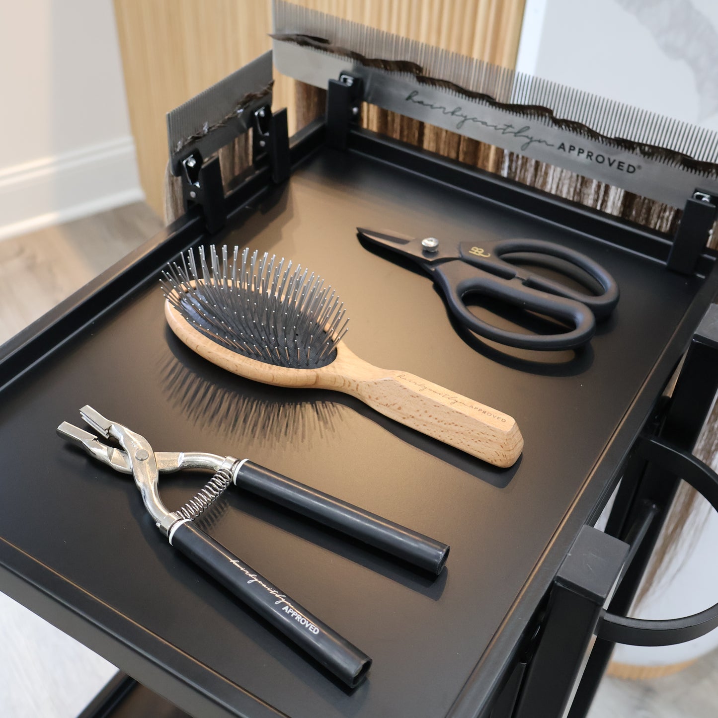 Set of hair styling tools including a brush, scissors, and pliers on a black surface.
